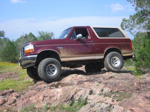 1996 Ford Bronco - A maroon 1996 Ford Bronco with a tan top navigates a rocky terrain under a blue sky.