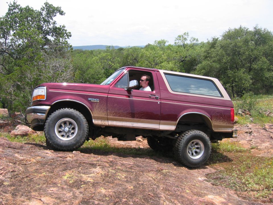 Maroon Ford Bronco with tan top driving on rocky terrain. Driver visible