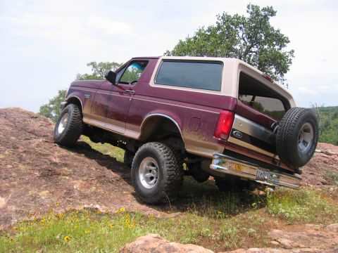 1996 Ford Bronco - A maroon Ford Bronco with a tan top navigates a rocky, off-road trail with yellow wildflowers.