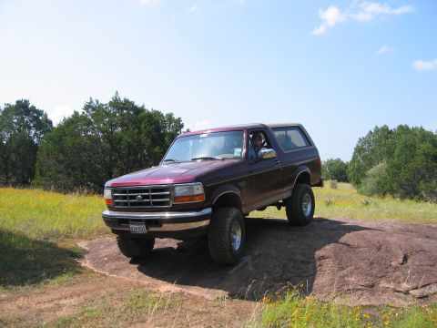 1996 Ford Bronco - This 1996 Ford Bronco is positioned on a large rock formation in a grassy, tree-lined area under a bright blue sky.