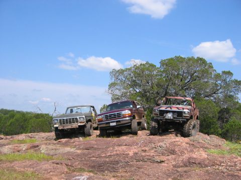 1996 Ford Bronco - Three off-road trucks, including a maroon 1996 Ford Bronco, are parked on a rocky hill under a blue sky.