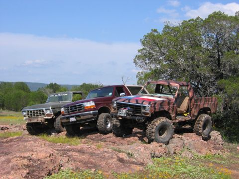 1996 Ford Bronco - Three off-road trucks are parked on a rocky terrain with trees and hills in the background.