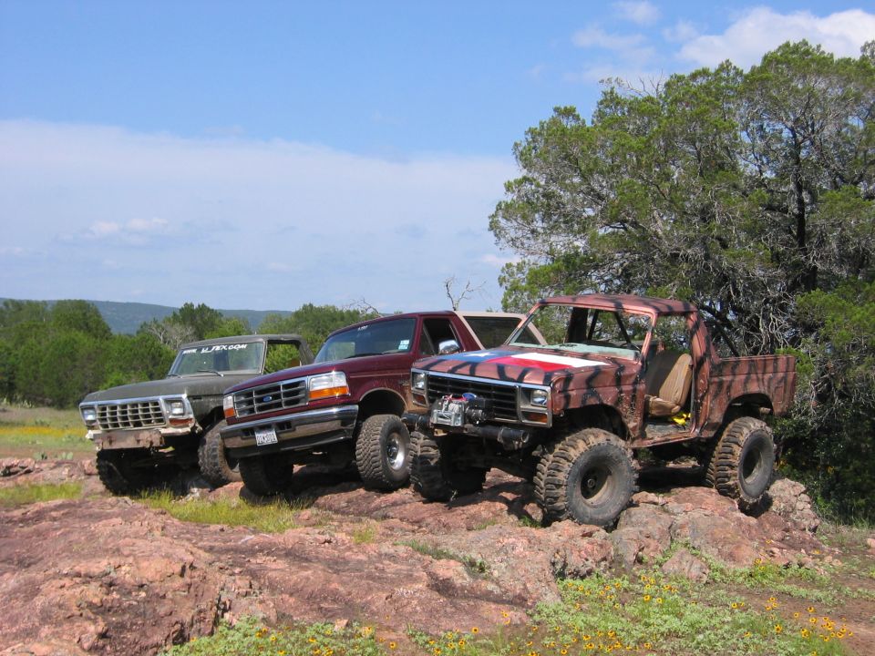 Three modified trucks parked on a rocky incline under a blue sky; the front truck has camouflage paint