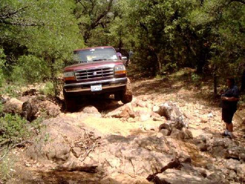 1996 Ford Bronco - A maroon Ford F-Series truck is shown off-roading on a rocky trail surrounded by trees.