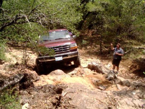 1996 Ford Bronco - A person directs a red Ford Bronco off-road through a rocky, wooded area.