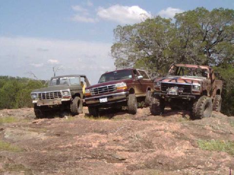 1996 Ford Bronco - Three modified trucks are parked on a rocky incline under a partly cloudy sky.