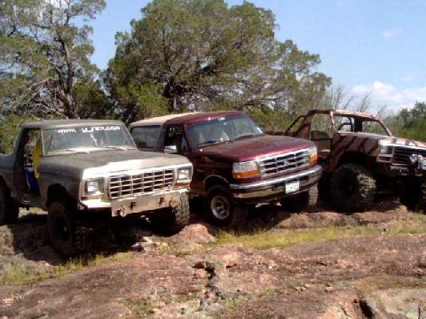 1996 Ford Bronco - Multiple trucks, including a maroon Ford Bronco, are parked on a rocky terrain under trees.