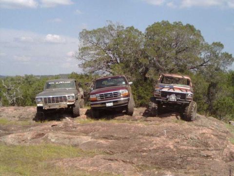 1996 Ford Bronco - Three modified trucks, including a Ford Bronco, are parked on a rocky hill under a partly cloudy sky.
