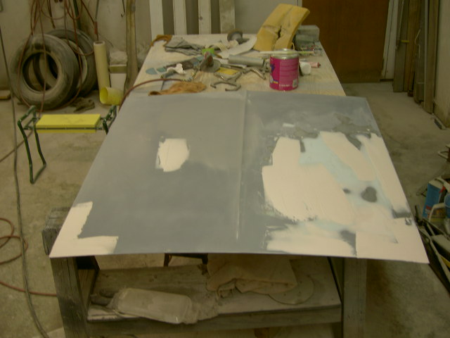 Car hood with gray primer and body filler patches, on a workbench