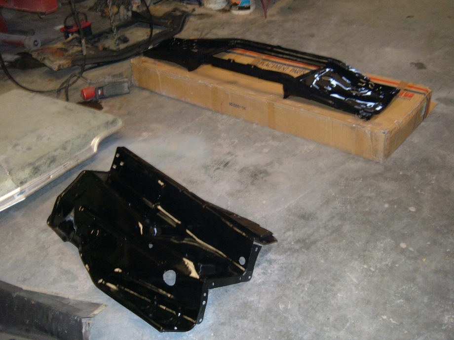 Two glossy black painted vehicle sheet-metal parts on a garage floor, one in a cardboard box.