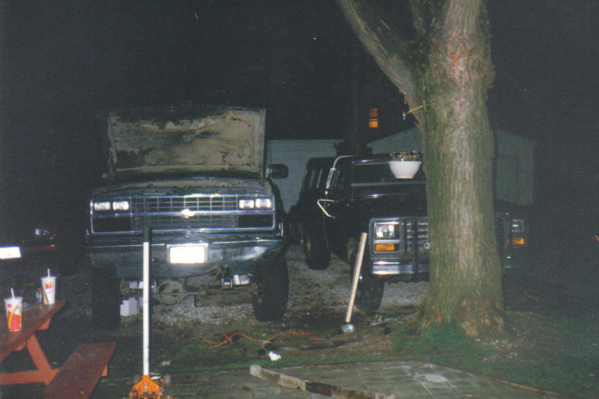 Dark GM truck with hood up at night beside another truck, tree, and picnic table with cups