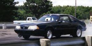 1989 Ford Mustang - A black 1989 Ford Mustang hatchback is seen on a drag strip, with smoke coming from the rear tire.