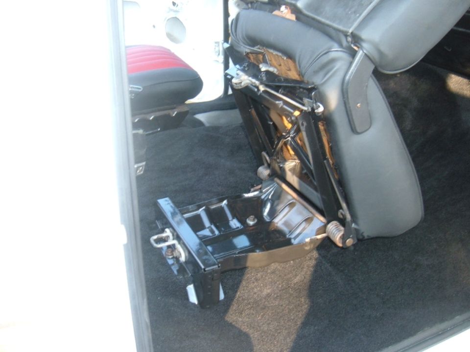 Car interior showing a removed seat base and black carpet. A red and black seat is visible in the background