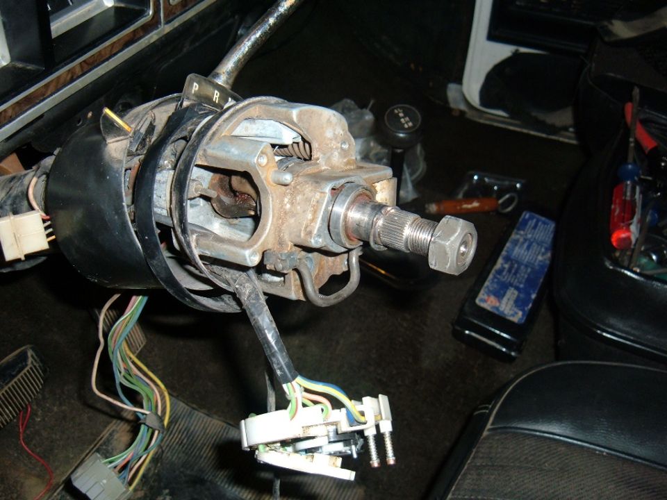 Vehicle's steering column assembly with wiring harnesses disconnected. Tools and a blue rag are on the floor