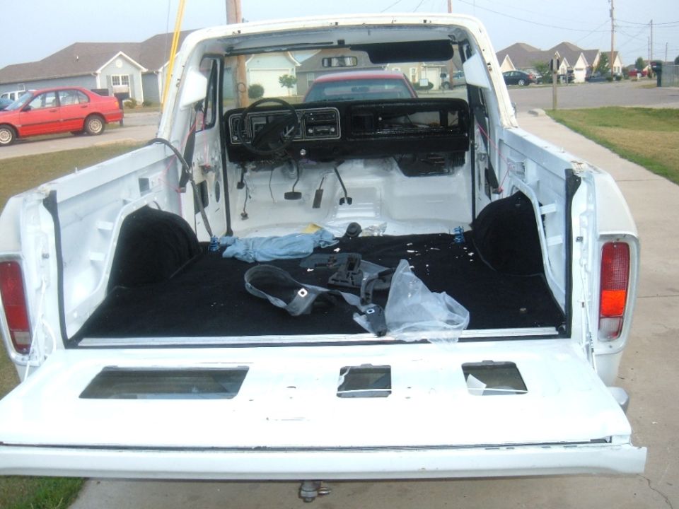 Disassembled interior and bed of a white pickup truck, possibly a 1979 Ford F-Series. Red car visible in background