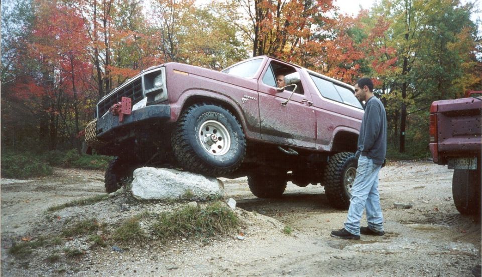 Lifted maroon Ford Bronco with front tire up on a boulder as a spotter watches, fall trees behind
