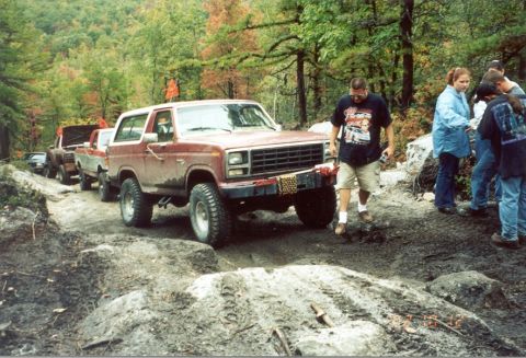 1980 Ford Bronco - A group of people and several vehicles are on a dirt trail in a wooded area.