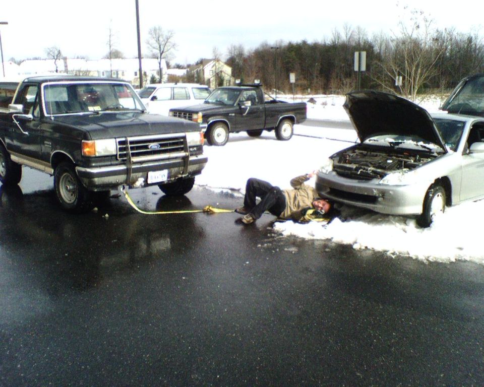 Ford Bronco with yellow tow strap pulling a silver car from snow while a man lies beside the car's bumper