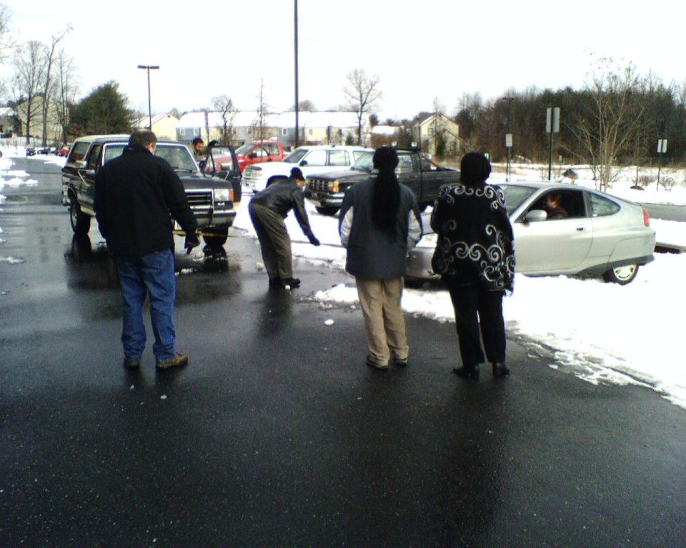 Dark Ford Bronco with yellow tow strap pulling a silver car in a snowy parking lot as people watch