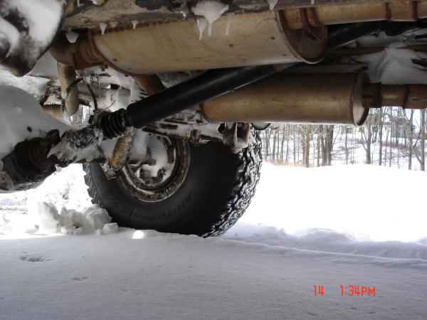 Underside of a 4x4 showing driveshaft, rear axle, muffler and all-terrain tire over snow