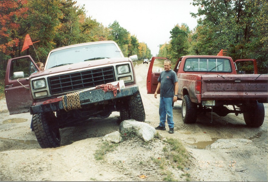 Lifted maroon Ford Bronco with door open and tow chain on the bumper beside a red Ford pickup