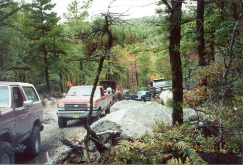1980 Ford Bronco - A maroon Ford Bronco is seen on a dirt road, with several other vehicles following behind it in a wooded area.