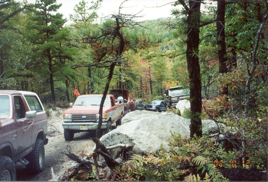 Convoy of 4x4 trucks staged on a rocky woods trail behind a boulder, maroon Bronco in foreground