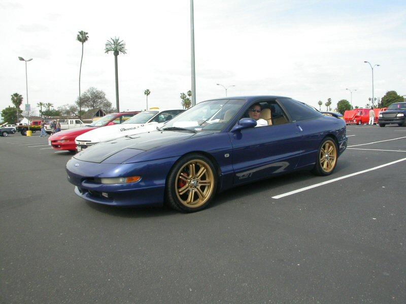 Blue 1994 Ford Probe GT with gold wheels parked in a lot, driver visible