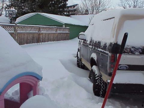1993 Ford Bronco - A dark blue Ford Bronco is partially buried in snow, with a red shovel resting against its rear.