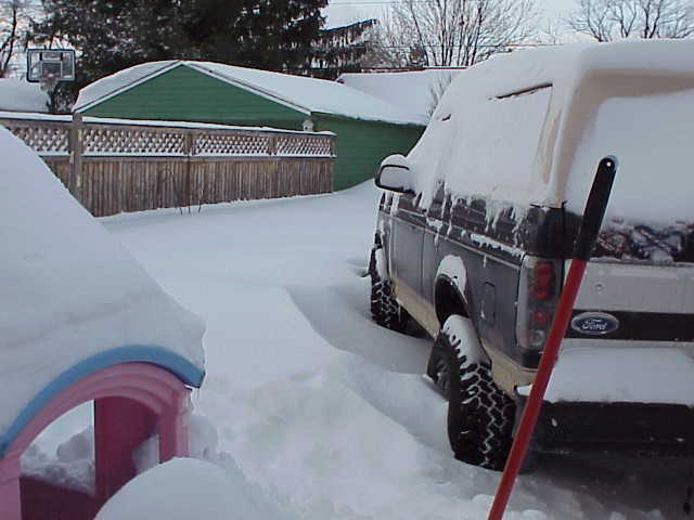 Dark blue Ford Bronco covered in snow, rear view, with a red shovel leaning against it