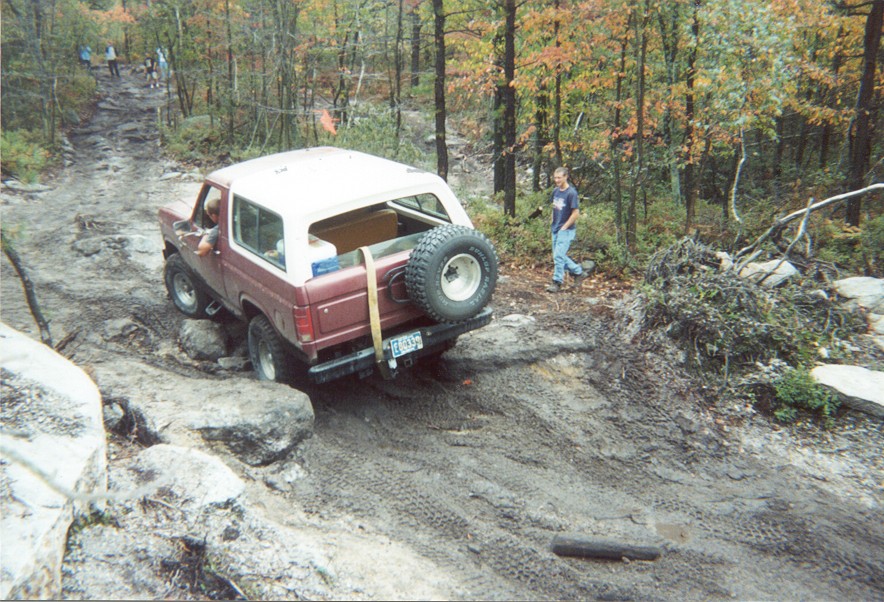 Rear view of a lifted maroon Bronco with tailgate-strapped spare tire climbing a rocky trail
