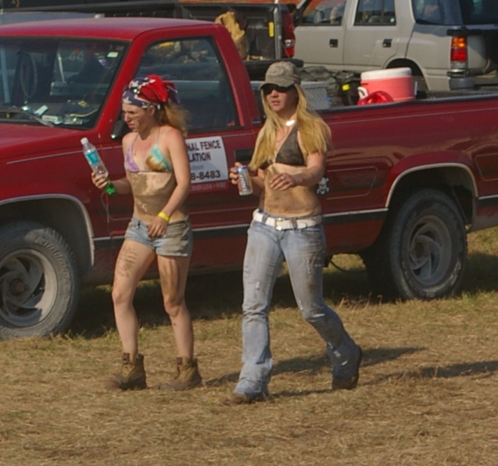 Two mud-covered spectators walking past a red work pickup at an outdoor mud event