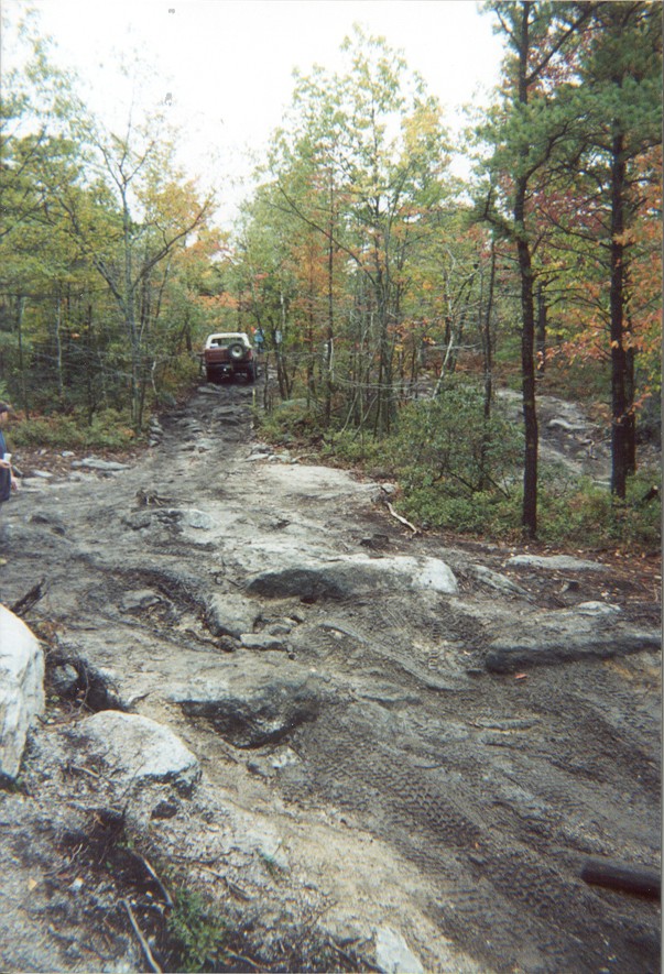 Rocky, tire-churned trail climb with a maroon SUV and rear spare tire cresting at the top