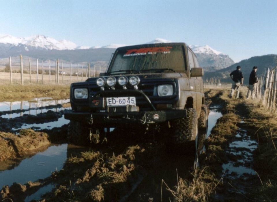 Muddy dark SUV with brush guard and roof lights stuck in muddy ruts, mountains behind