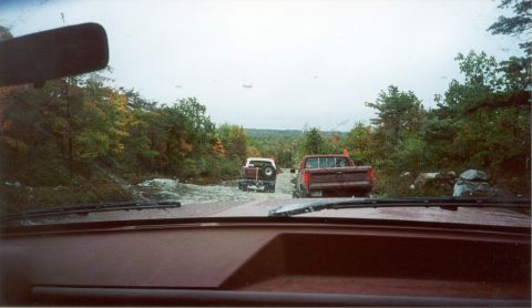 1980 Ford Bronco - Two trucks travel down a dirt road during autumn with trees lining the path.