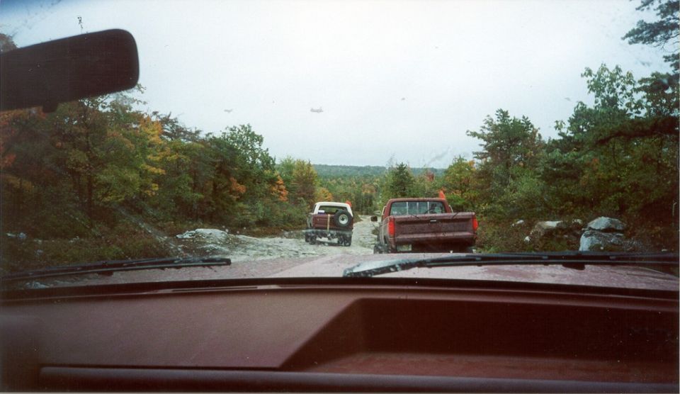 Driver's-eye windshield view of a white-topped SUV and red pickup ahead on a rocky trail
