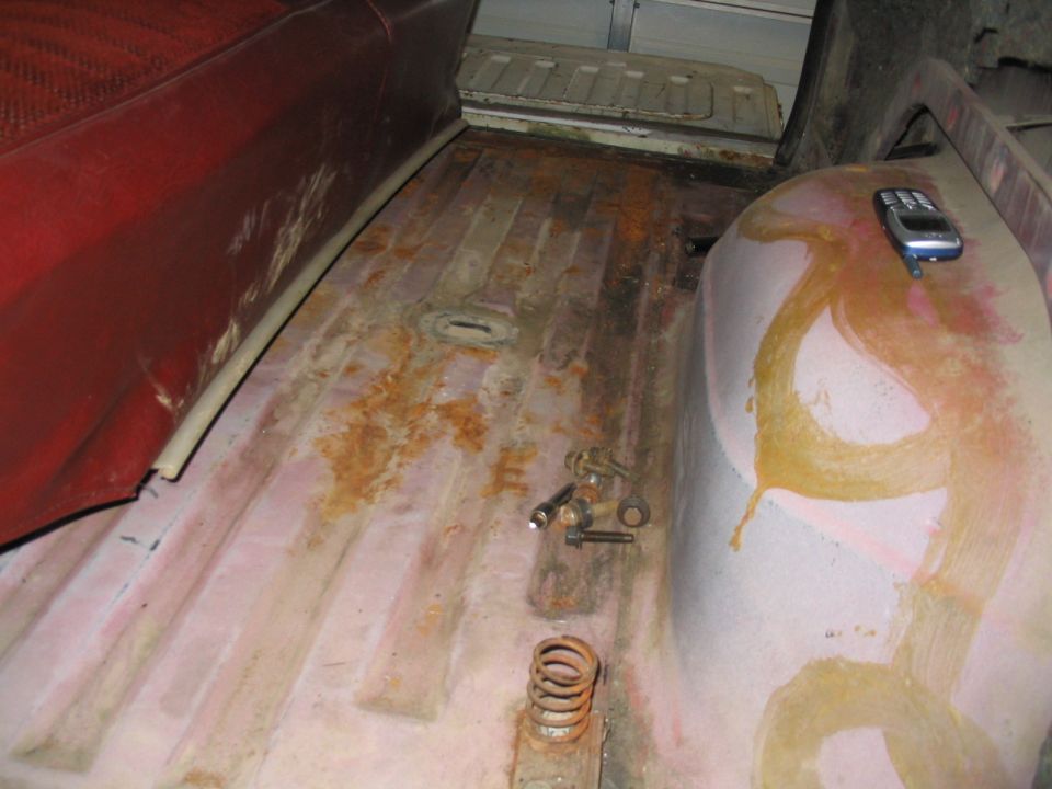 Stripped Bronco interior floor pan with surface rust, coil spring and bolts, red seat at left, phone on wheel well
