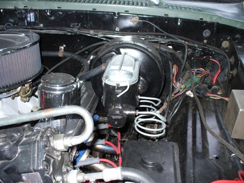 1978 Ford Bronco - Overhead engine-bay view of a black brake booster and master cylinder with coiled steel brake lines mounted on a black firewall, with a col…