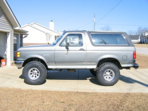 1989 Ford Bronco - A lifted 1989 Ford Bronco is shown from the side, parked on a paved surface in front of a residential garage.