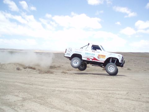 1990 Ford Bronco - White lifted off-road race truck catching air over a desert dirt jump, kicking up dust under a blue cloudy sky.