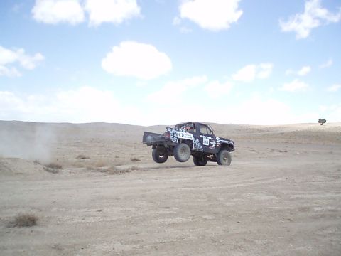 1990 Ford Bronco - Dark-colored off-road race truck airborne over a desert course, trailing a cloud of dust under a bright sky.