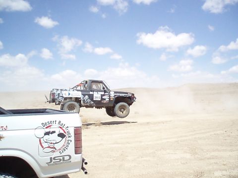 1990 Ford Bronco - Black number 301 off-road race truck airborne in the desert, viewed past the bed of a white Desert Rat tow truck.