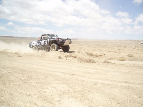 1990 Ford Bronco - Black off-road race truck sliding through a dusty desert corner, spraying dirt as it powers around the berm.