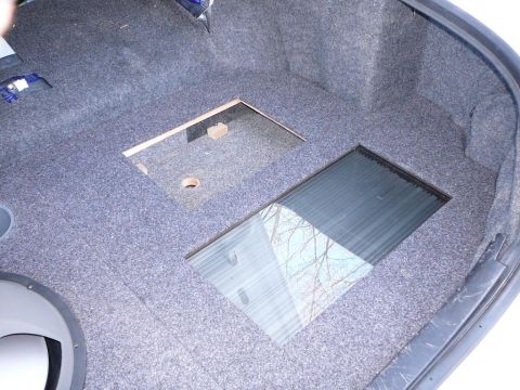 1997 Mercury Grand Marquis - Trunk interior of a sedan lined with dark gray carpet, showing a custom subwoofer enclosure build with a rectangular MDF cutout, a small mo…