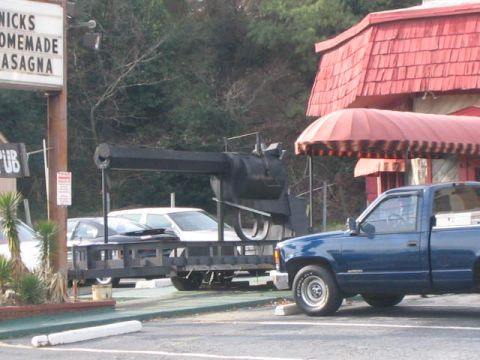 1979 Ford Bronco - A large novelty revolver is displayed on a trailer, with a blue pickup truck parked in front of a building.