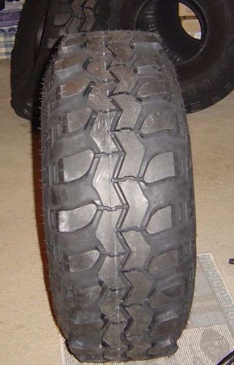 1986 Ford Bronco - Close-up of the tread face of a used mud-terrain tire standing upright on carpet indoors, showing chunky staggered lugs, siped shoulder blo…