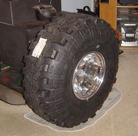 1986 Ford Bronco - Aggressive mud-terrain tire mounted on a polished aluminum deep-dish wheel, standing on carpet indoors beside a wood stove. Tall square-sho…