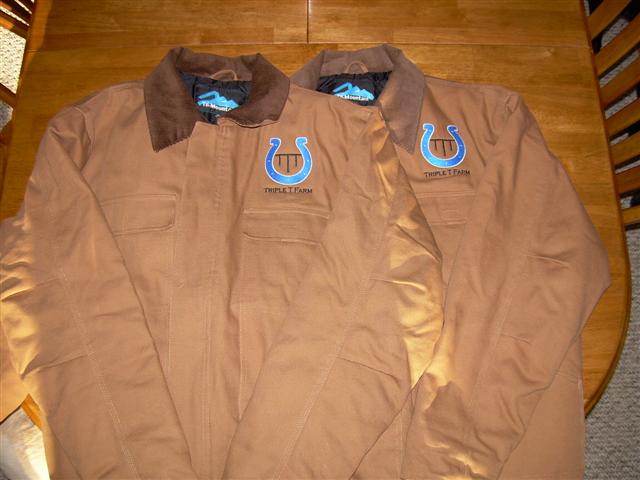 two tan work jackets with blue horseshoe and 'TRIPLE T FARM' logo on them, laid on a wooden table