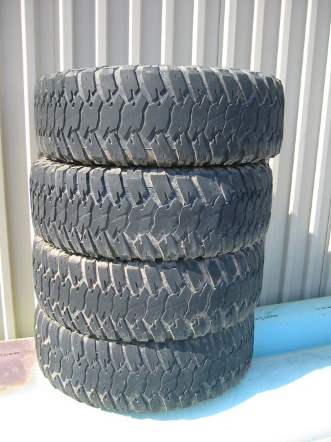 1966 Ford Bronco - Four off-road tires stacked up against a corrugated metal wall.