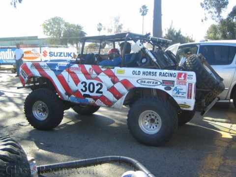 1990 Ford Bronco - Side profile of a patriotic stars-and-stripes early-Bronco race truck number 302 with full roll cage and rear spare tire.